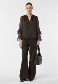 Blouse noire en tissu transparent avec manches à volants, col en V et détail à cordon ; associée à un pantalon évasé marron foncé. Le mannequin tient un sac fourre-tout marron.