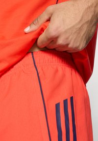 Élénk vörös sportpóló kék részletekkel. Közeli kép elasztikus derékú nadrágról, amelyen három sötétkék csík található, és puha tapintású.