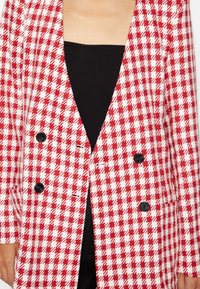Červeno-bílý kostkovaný blazer, dvouřadý design se dvěma černými knoflíky, strukturovaná tkanina a hluboký V-výstřih.
