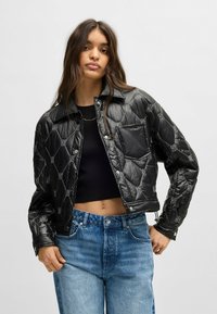 Black quilted jacket with shiny finish, snap buttons, and graphic accents. Features a cropped design and a single chest pocket.
