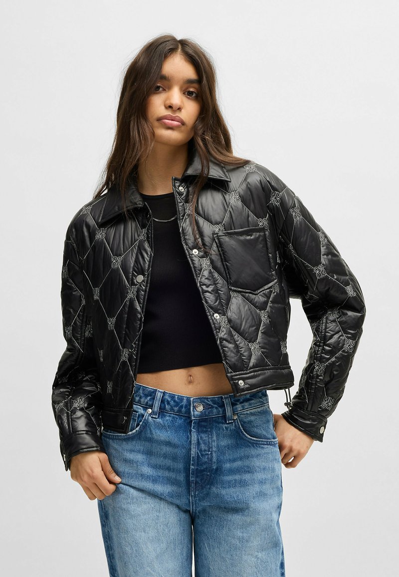 Black quilted jacket with shiny finish, snap buttons, and graphic accents. Features a cropped design and a single chest pocket.