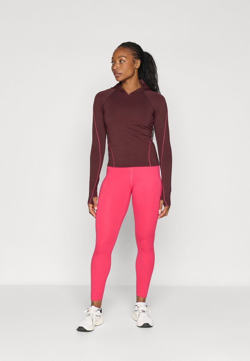 Bordeauxfarbenes Langarm-Sportoberteil mit pinken Akzenten, kombiniert mit leuchtend pinken Leggings. Das Model trägt weiße Sneakers. Glattes Material, figurnaher Schnitt.