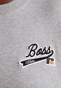 Tissu gris avec un écusson cousu portant l'inscription "Boss Athletic" en noir et blanc, avec une petite lettre R stylisée en noir et orange.