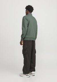 Sudadera verde con alto cuello, combinada con pantalones cargo negros holgados y zapatillas blancas. El conjunto presenta una textura suave y un diseño minimalista.