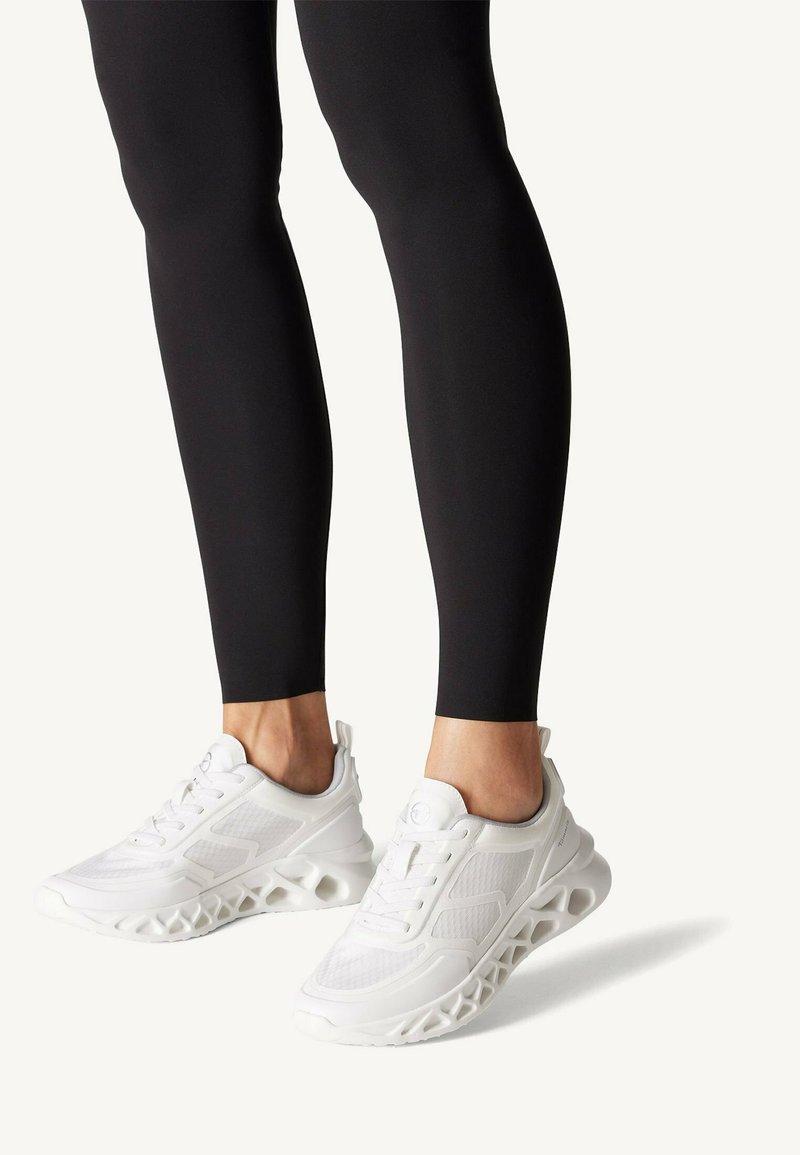 Valged spordijalatsid, millel on hingav võrkkangas pealisosa ja ainulaadne lõigetega talvekujundus. Kanda istuvate mustade retuusidega, rõhutades kingade detaile.