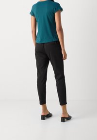 Teal kortärmad topp med spetsdetaljer, i kombination med svarta slim-fit byxor. Individen bär svarta slide-sandaler.