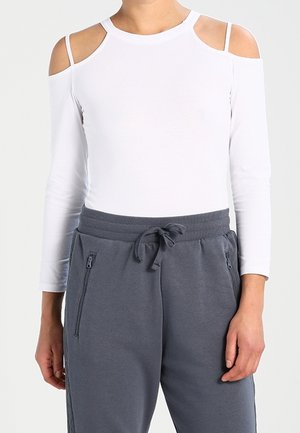 Weißes Langarmoberteil mit Auschnitten an den Schultern, kombiniert mit grauen Jogginghosen mit Reißverschluss-Taschen und verstellbarem Kordelzug in der Taille.