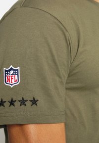 Olivově zelené tričko vyrobené z bavlny. Má vyšívané logo NFL a černé hvězdy podél rukávu. Hladká textura, design s krátkým rukávem.