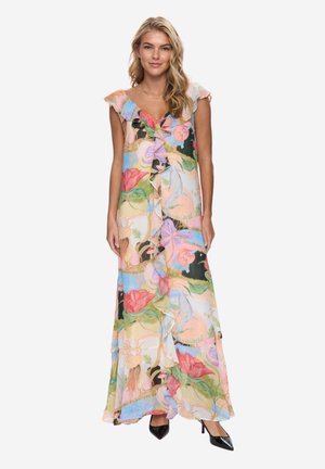 Kobieta stojąca, ubrana w bezrękawnikową, falbanową sukienkę maxi w pastelowe kwiaty oraz czarne czółenka z ostrym noskiem.