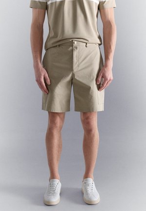 Mann trägt beige Cargo-Shorts, ein passendes beige kurzärmeliges Hemd und weiße Sneaker, steht vor einem einfarbigen Hintergrund.