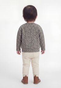 Mazs bērns stāv ar muguru pret skatītāju, tērpies pelēkā ziedu raksta krekliņā ar garām piedurknēm, krēmkrāsas ribotas biksēs un brūnu zābaciņos uz baltas fona.