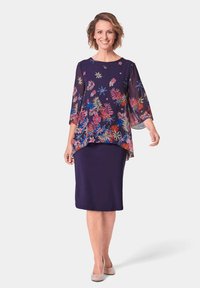 Bluse mit Blumenmuster in Marineblau mit Dreiviertelärmeln, über einem taillierten marineblauen Rock getragen. Glatte Stoffstruktur, dezente Glitzerakzente am Ausschnitt.