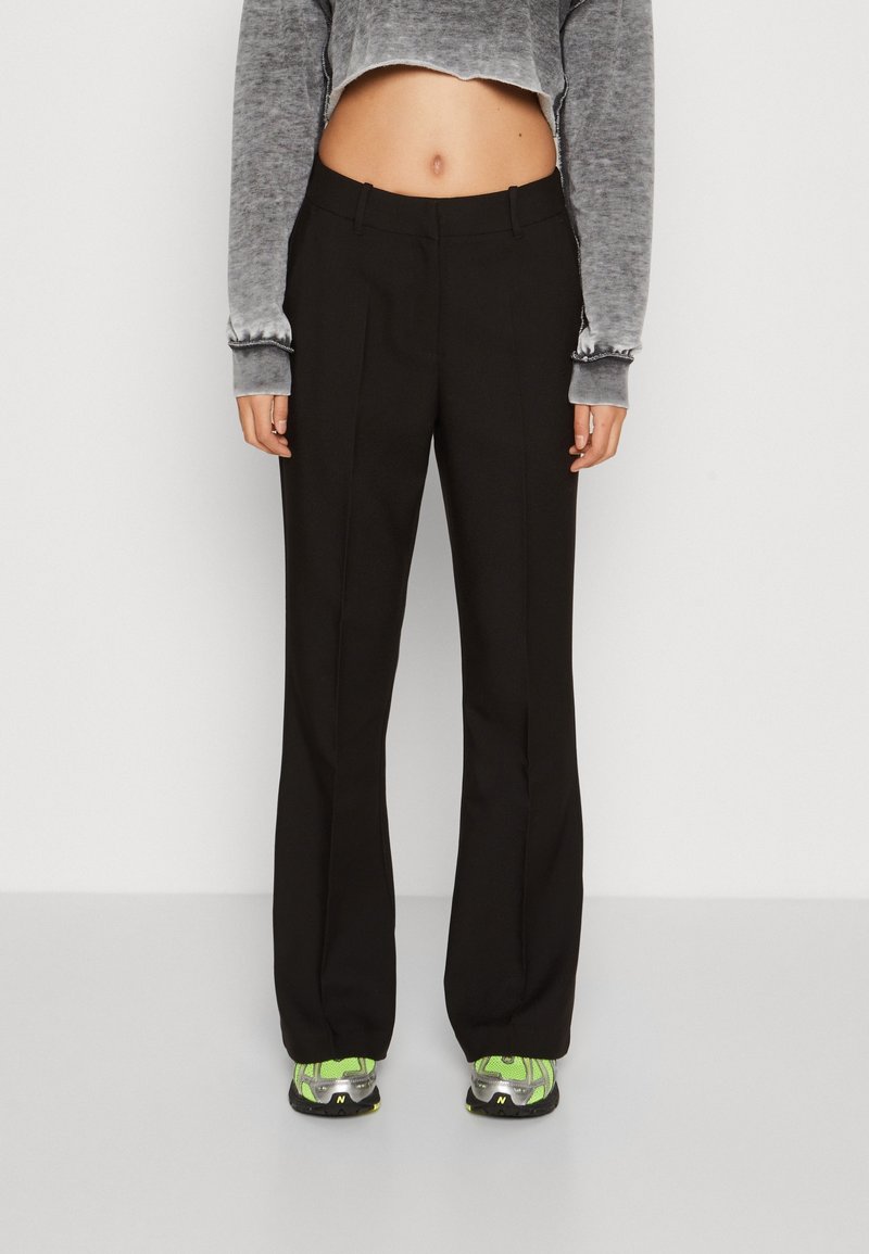 Personne portant un pantalon droit noir, un crop top gris à manches longues et des baskets vertes et grises, debout devant un fond uni.