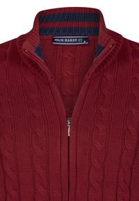 Cárdigan de punto trenzado rojo con cierre frontal, con un cuello a rayas en azul marino. Hecho de lana gruesa, que ofrece textura y calidez.