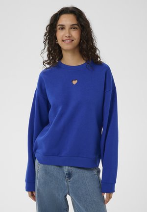 Mladá žena s kučeravými vlasmi, oblečená v modrom svetri s malým srdcovým výrezom a svetlomodrých džínsoch, usmievajúca sa na jednoduchom pozadí.