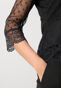 Blouse noire avec manches en maille à pois semi-transparente et bordure en dentelle délicate. Le corps est noir uni avec une coupe ajustée.