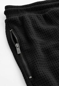 Pantalons en tissu noir texturé avec une poche zippée métallique visible et un détail de taille élastique sur fond blanc.