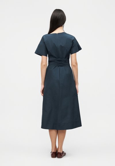 Γυναίκα με μακριά σκούρα μαλλιά, ντυμένη με σκούρο μπλε φορέμα μίντι με κοντά μανίκια, εφαρμοστή μέση και καφέ επίπεδα παπούτσια, εμφανιζόμενη από πίσω.