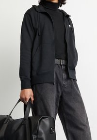 Svart zip-up hoodie med en framficka, kombinerad med en svart polotröja och mörkgrå jeans. Modellen håller en svart duffelväska.