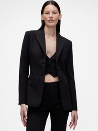 Blazer noir aux épaules structurées, avec deux poches avant et un revers cranté. Porté sur un gilet court noir assorti et un pantalon tailleur.
