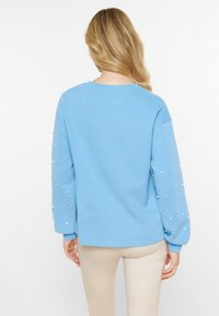 Jasnoniebieski sweter z okrągłym dekoltem i długimi bufkami, ozdobiony małymi białymi perłami. Gładka tekstura materiału, codzienny krój.