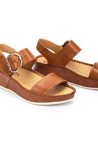 Chanclas de cuero marrón con un hebilla metálica redonda, diseño de punta abierta, detalles cosidos a lo largo de los bordes y una suela de color claro.