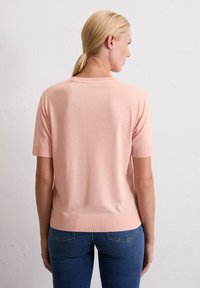 Haut rose clair à manches courtes avec un col rond, des poignets et un ourlet côtelés ; texture lisse et silhouette décontractée. Jeans visibles en bas.