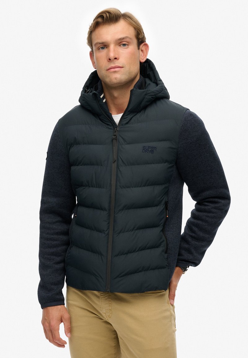 Giacca imbottita blu navy con cappuccio aderente e maniche in lana blu, dotata di una zip sul davanti e dettagli con il logo. Contrasti di texture tra i materiali.