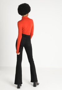 Röd polokrage med långa ärmar, parat med högmidjade svarta utsvängda jeans. Svarta klackstövlar. Neutral bakgrund.