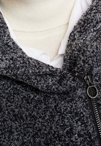 Veste grise texturée avec une finition duveteuse, fermeture éclair et capuche. Portée sur une chemise blanche et un col roulé crème.