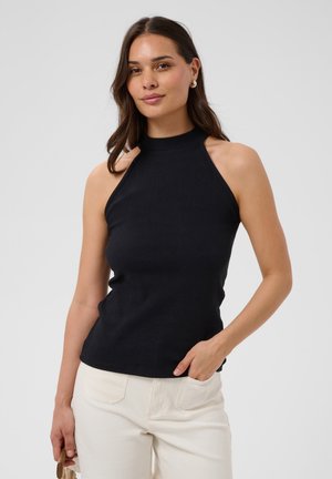 Femme aux longs cheveux foncés, portant un haut noir sans manches et un pantalon blanc, debout avec une main dans la poche devant un fond uni.