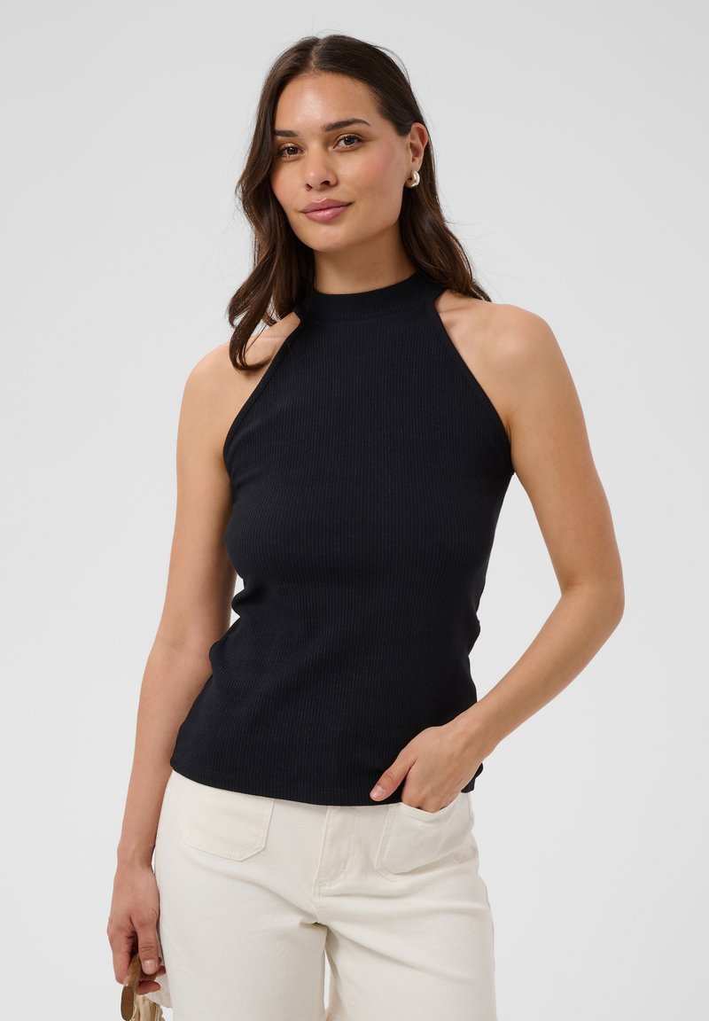 Femme aux longs cheveux foncés, portant un haut noir sans manches et un pantalon blanc, debout avec une main dans la poche devant un fond uni.