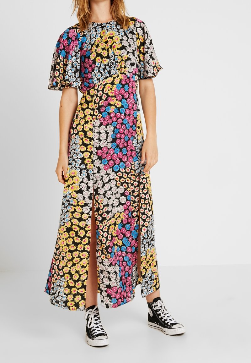 Frau in einem bunten, blumigen Patchwork-Maxi-Kleid mit kurzen Ärmeln und Schlitzen vorne, kombiniert mit schwarzen High-Top-Sneakern.