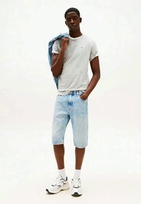 Grå bomullströja, ljusblå denimshorts och vita sneakers. Modellen håller en denimjacka; avslappnad passform med en enkel design.