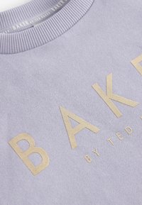 Svetlo levanduľový sveter s rebrovaným výstrihom, na ktorom je výrazný zlatý nápis "BAKER" a menší text "by TED BAKER" v hladkej textúre.