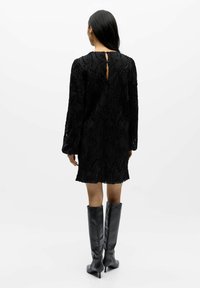 Schwarzes Kleid aus strukturiertem Stoff mit einem abstrakten Muster; halb-lange Ärmel; Schlitz im Rücken; getragen mit hohen schwarzen Stiefeln.