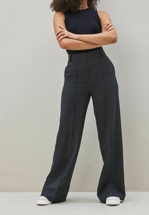 Kvinde iført sort ærmeløs top, højtaljede viddebukser i mørkegrå med nålestriber og hvide sneakers, stående med armene over kors.