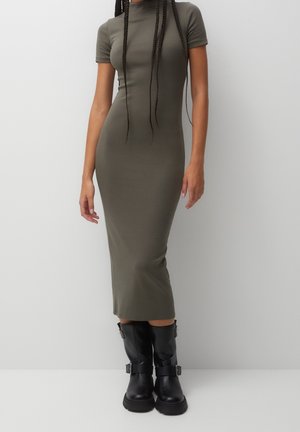Woman wearing an olive green, short-sleeve, ankle-length fitted dress with black mid-calf buckle boots, standing against a plain white background.