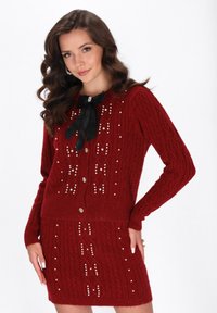 Punainen neulepaita ja hamekokonaisuus, jossa on kaapelikuvio, helmet ja musta rusetti kauluksessa, sekä napituksella varustettu neulepaita.