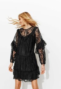 Robe en dentelle noire avec manches longues transparentes, encolure volantée et jupe à volants. Ouverture en forme de goutte d'eau à l'avant, texture douce et motif floral.