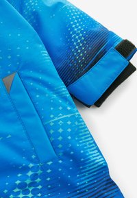 Veste bleue avec une texture brillante, présentant un motif géométrique et une poche latérale. Les accents incluent du tissu noir au niveau des poignets.