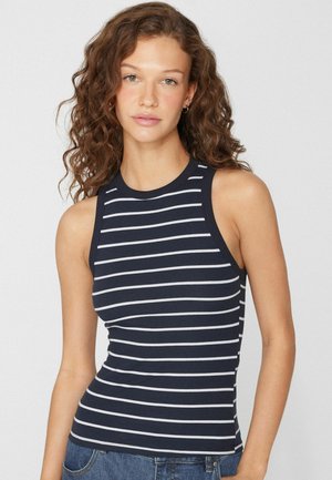 Femme aux cheveux bouclés portant un haut côtelé sans manches bleu marine avec des rayures horizontales blanches, associé à un jean bleu.