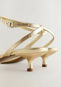 Gouden metallic hoge sandaaltjes met kruislings banden, lage kittenhakken en een gladde afwerking. Gespsluiting op de verstelbare enkelband.