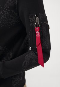 Černá mikina se subtilními vzory peří, kapsou na zip na manžetě a červenou tkaninou s logem na stříbrném kovovém prvku.