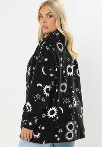 Mustap fleecetakki, jossa on taivaallinen kuvio valkoisista kuuista, auringoista ja tähdistä. Pehmeä rakenne, löysä istuvuus ja korkea kaulussuunnittelu.