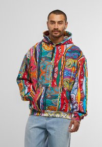 Mann in einem bunten, gemusterten Hoodie und hellblauen Jeans, steht vor einem schlichten, hellgrauen Hintergrund.