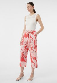 Weiße, gerippte Tanktop kombiniert mit lockeren, korallenfarbenen Hosen, die ein weißes Blumenmuster aufweisen. Das Model steht mit den Händen in den Taschen und trägt Sandalen mit Riemchen.