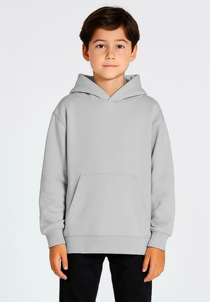 Jongen staat rechtop met zijn gezicht naar voren, gekleed in een lichtgrijze pull-over hoodie en zwarte broek, tegen een effen witte achtergrond.