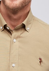 Beige színű, rövid ujjú ing, fehér gombokkal, gallérral, és részletes hímzett logóval a bal mellrészen.