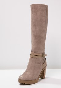 Bota de ante taupe hasta la rodilla con un tacón grueso, que presenta una correa decorativa con hebilla y una suela de plataforma texturizada. Acabado suave.